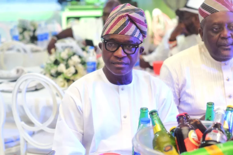 Man in striped cap and white tunic sits at a reception table beside drinks.
