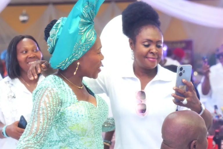 Two women in turquoise and white outfits laugh while taking a phone photo.