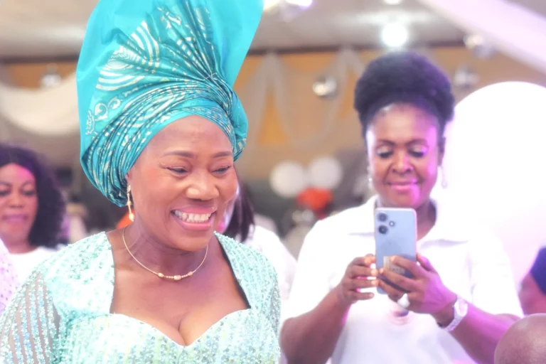 Woman in turquoise headwrap laughs as another in white films her on a phone.