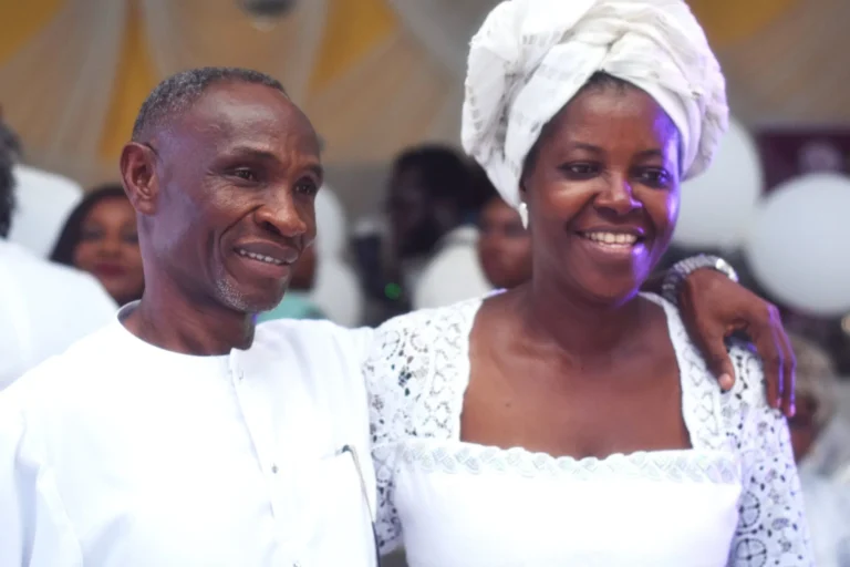 Smiling couple in white outfits pose with arms around each other at a party.