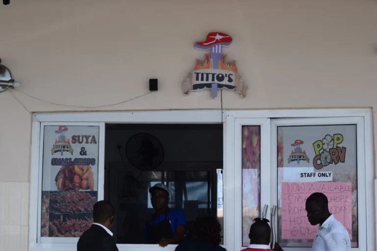 Exterior of Titto's takeaway with Suya, Small Chops and Popcorn posters, customers gathered at the hatch.