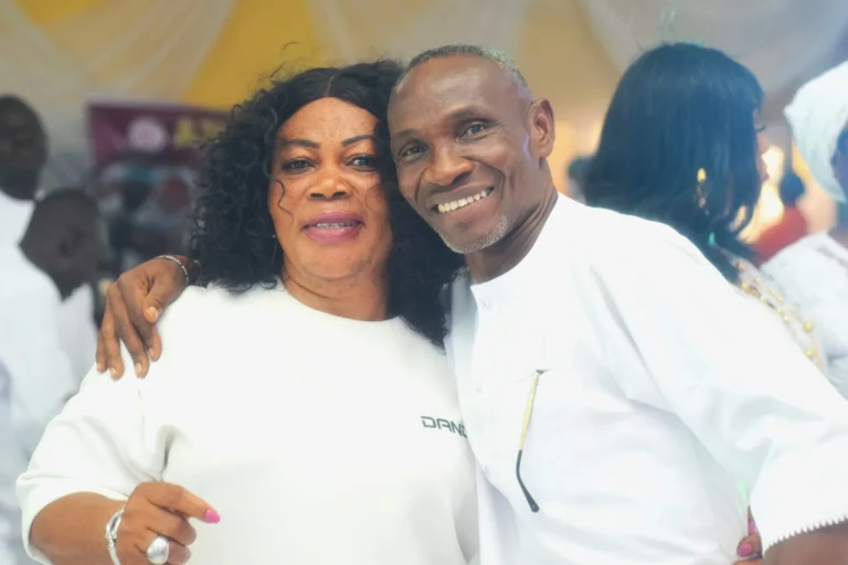 Smiling couple in white embrace warmly at a crowded indoor celebration.