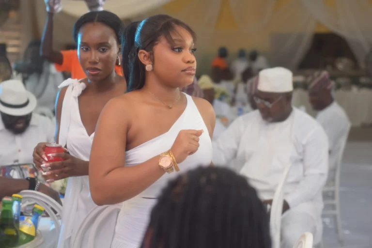 Two young women in white dresses stand at a guest reception, one mid-glance.