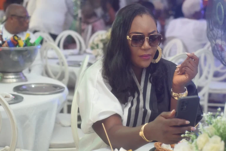Woman in sunglasses and striped dress checks her phone at a banquet table.