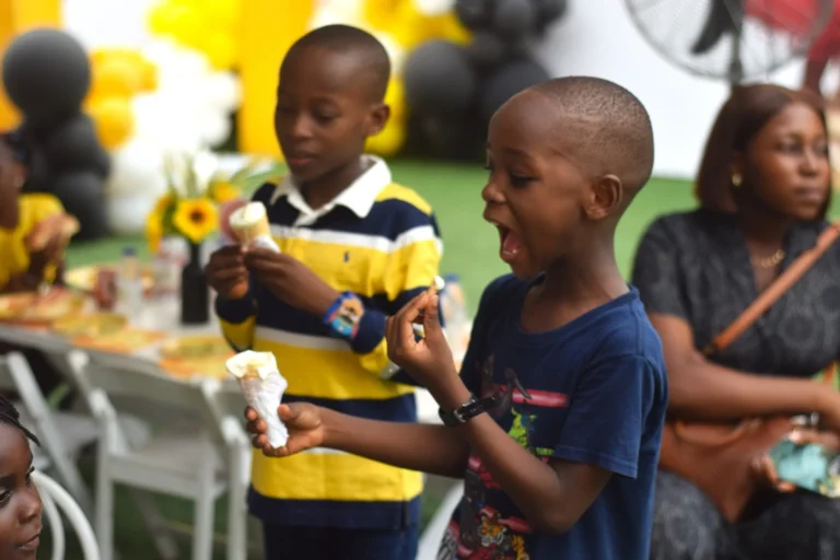 Two boys eating popcorn from striped cones at an outdoor party, one looking surprised.