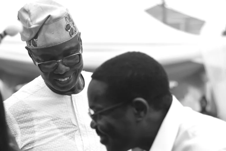 Black-and-white portrait of two men laughing together, one wearing a traditional Yoruba cap.