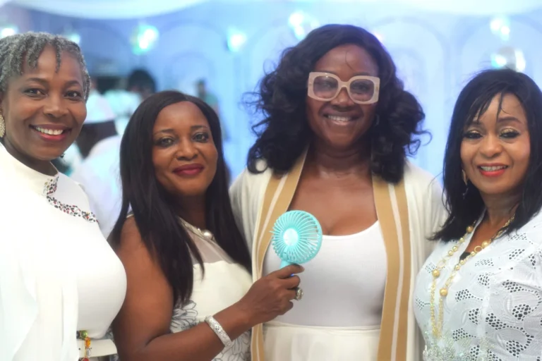 Four women in white outfits pose closely, one holding a small turquoise fan.