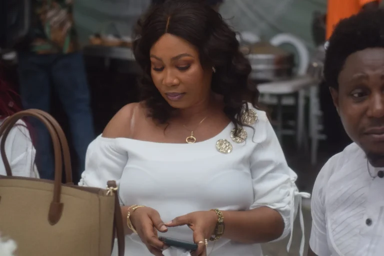 Woman in white off-shoulder dress glances at her phone holding a tan handbag.