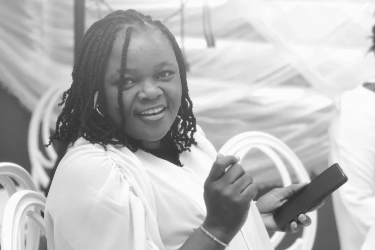 Smiling young woman with locs turns to camera holding a phone, in black and white.
