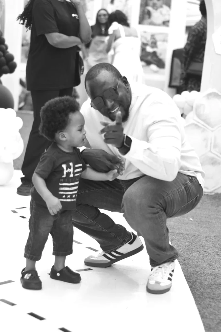 Black-and-white image of a man crouching to talk to a toddler boy on a walkway.