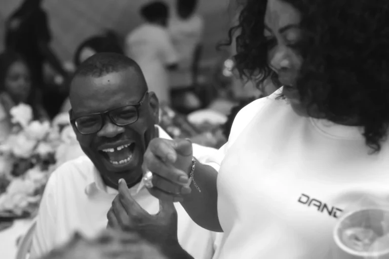 Man in glasses laughs heartily as a woman gestures beside him in black and white.