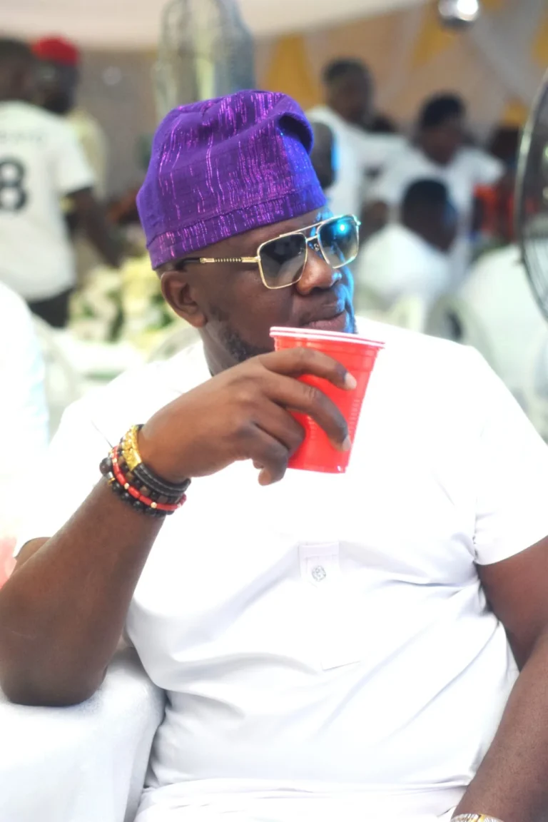 Man in purple cap and sunglasses sips from a red cup at a marquee event.