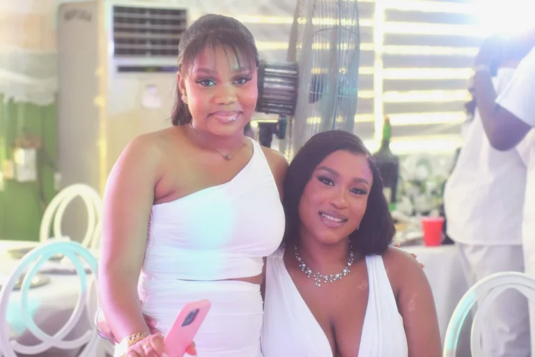 Two young women in white dresses pose together smiling at a reception.