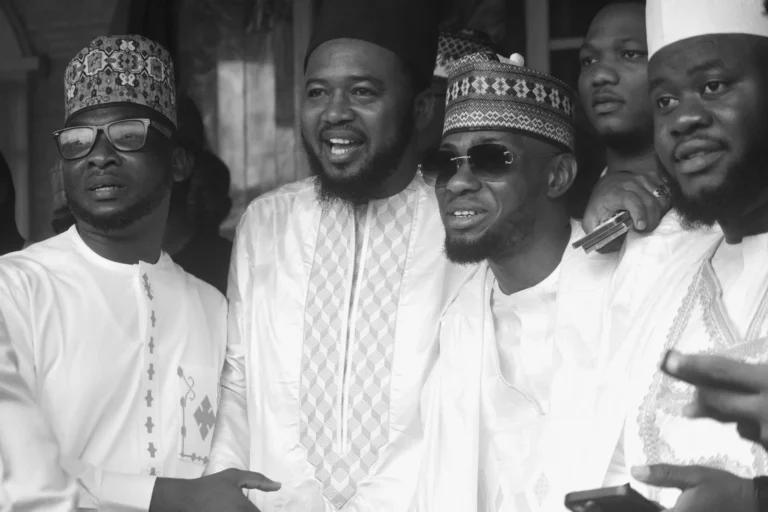 Four men in white agbada and patterned caps smiling for a group portrait.