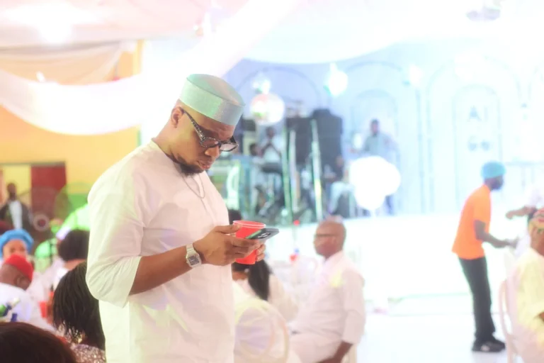Man in pale cap and white tunic checks his phone at a brightly lit reception.