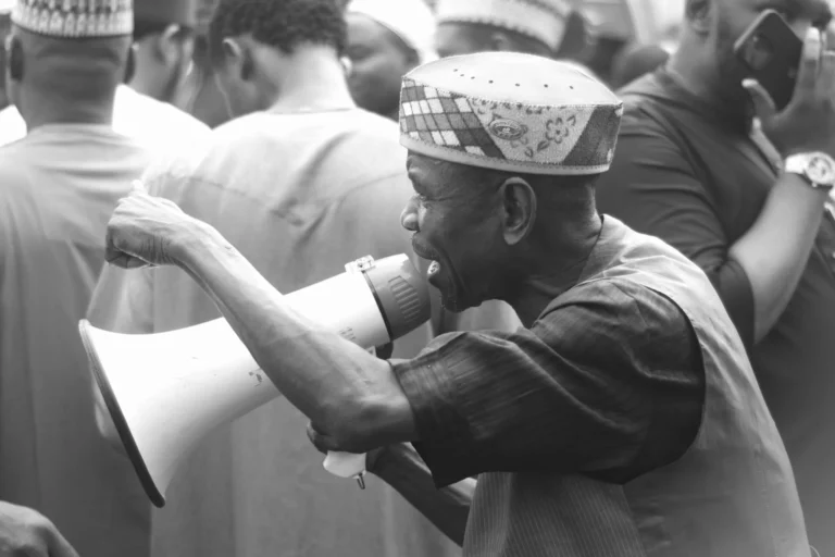 Elderly man in patterned cap shouting through a white megaphone amid a crowd.