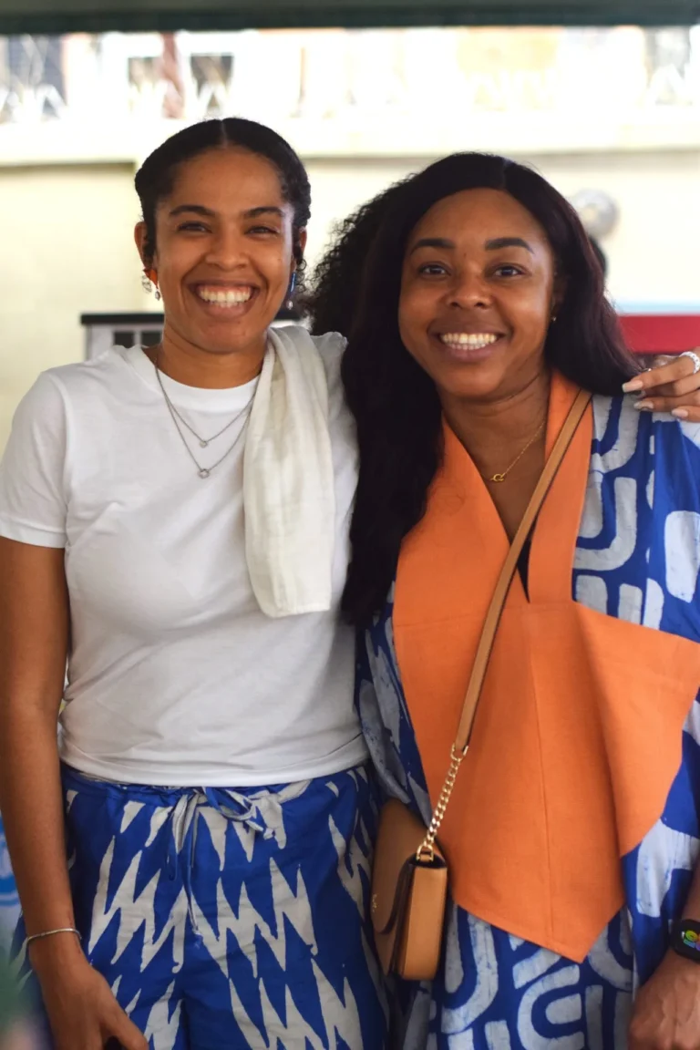 Two women embrace and beam for the camera, one in white tee and one in orange and blue.