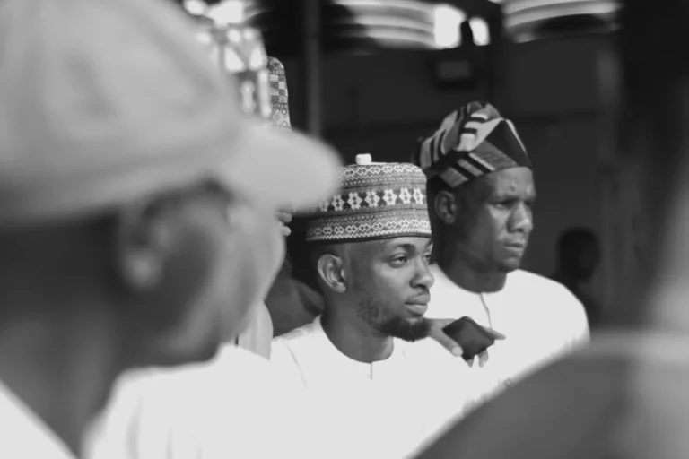 Young man in patterned cap stands among others, looking pensively to the side.