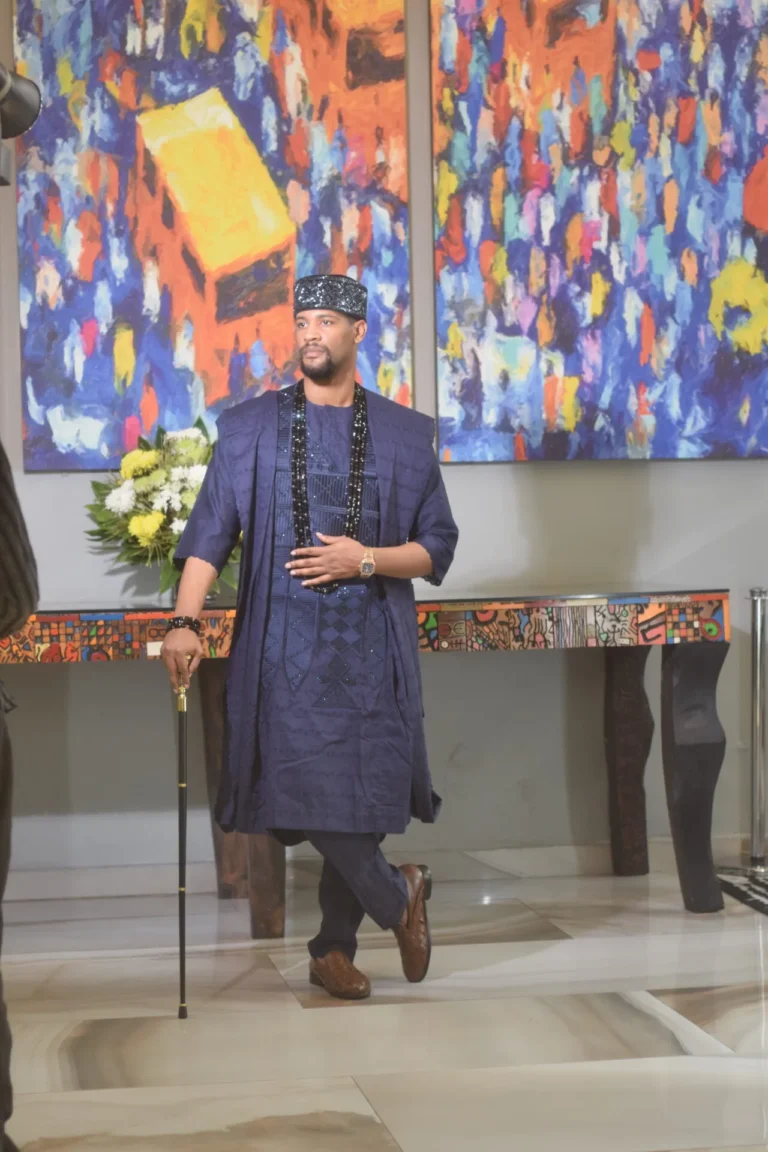 Man in blue agbada and beaded cap poses with a cane before abstract paintings.