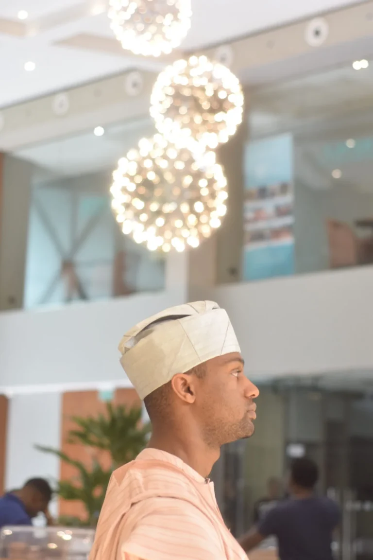 Man in cream cap and pink robe stands in profile beneath cluster of pendant lights.