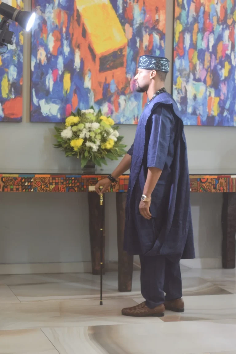 Man in dark blue agbada stands in profile beside floral arrangement and bright artwork.