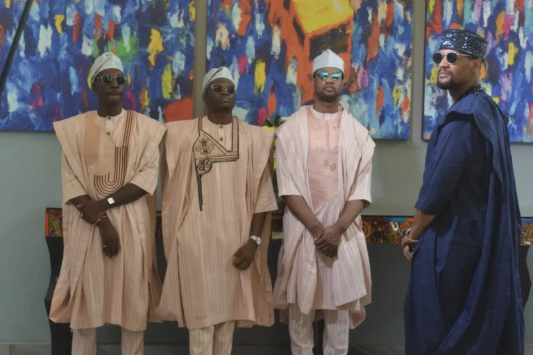 Four men in pastel agbada and caps stand before a colourful abstract painting.