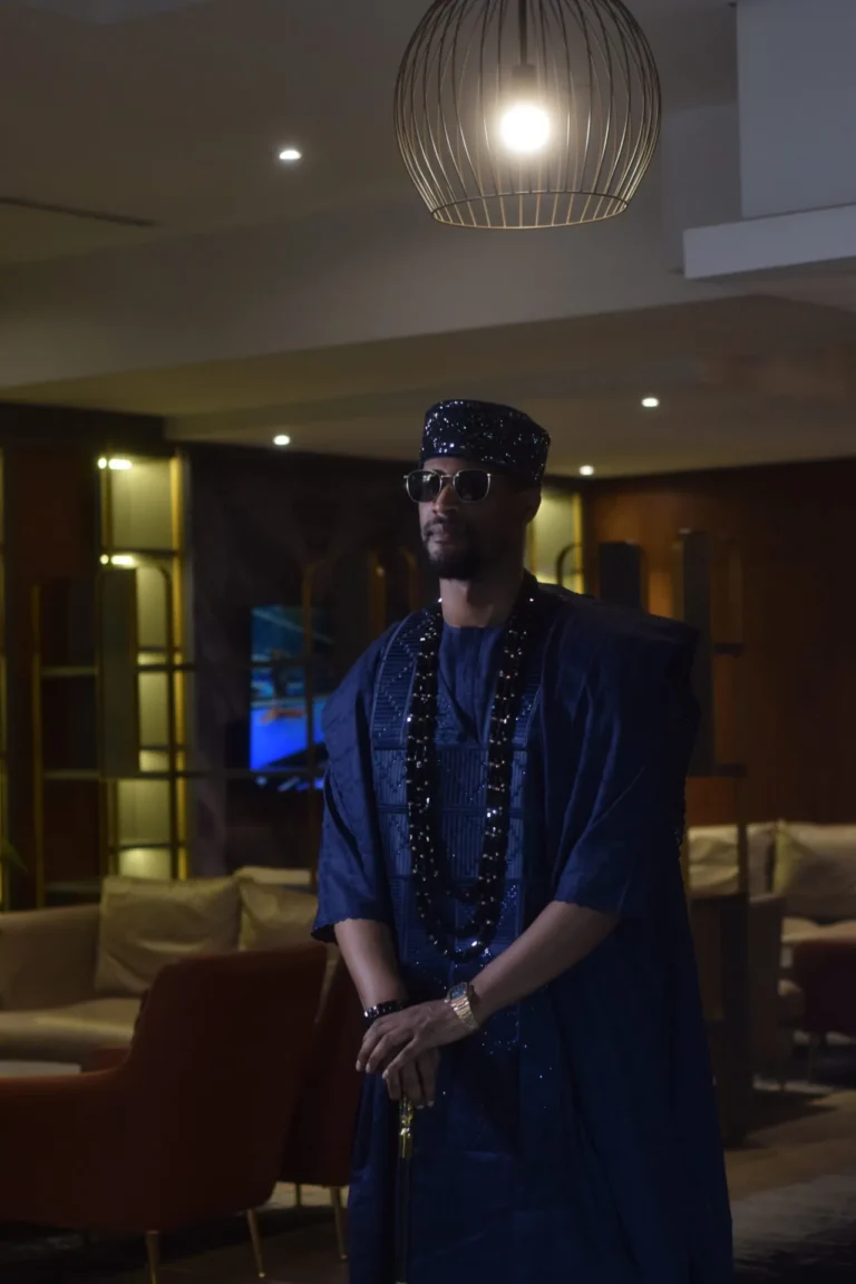 Man in navy agbada and sequinned cap stands beneath a spherical pendant light.