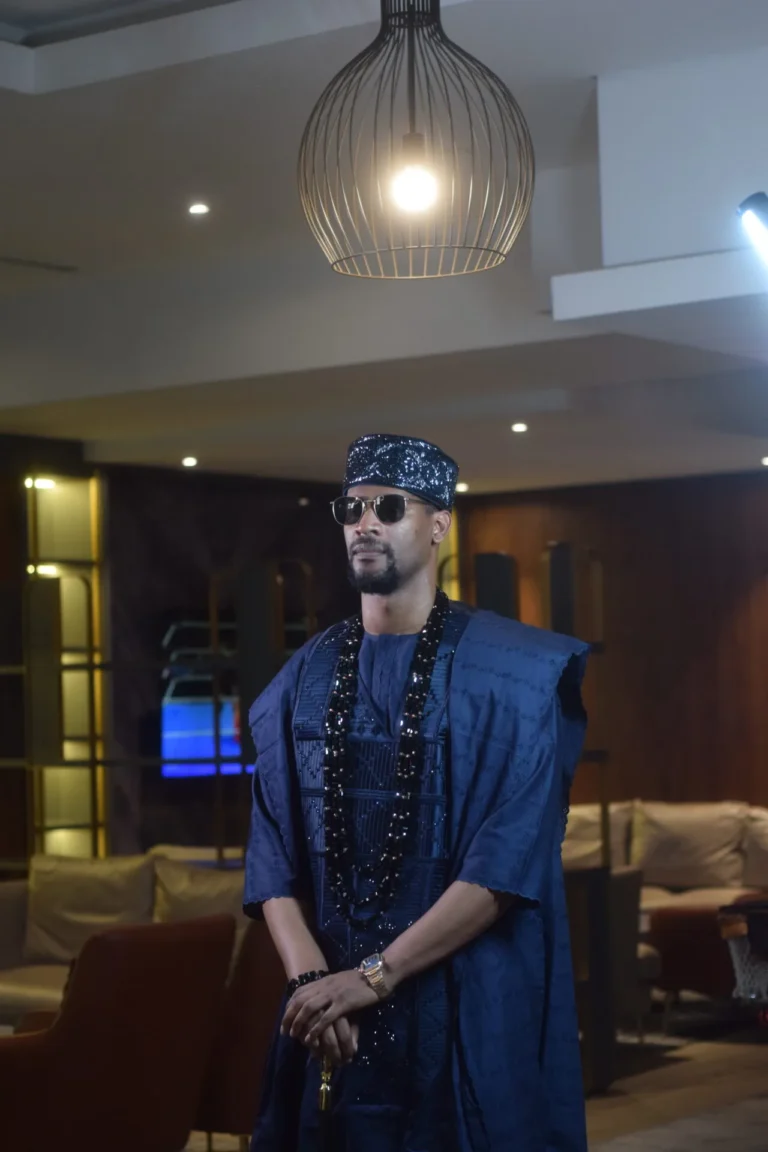 Man in navy agbada and beaded cap poses indoors under a teardrop pendant lamp.