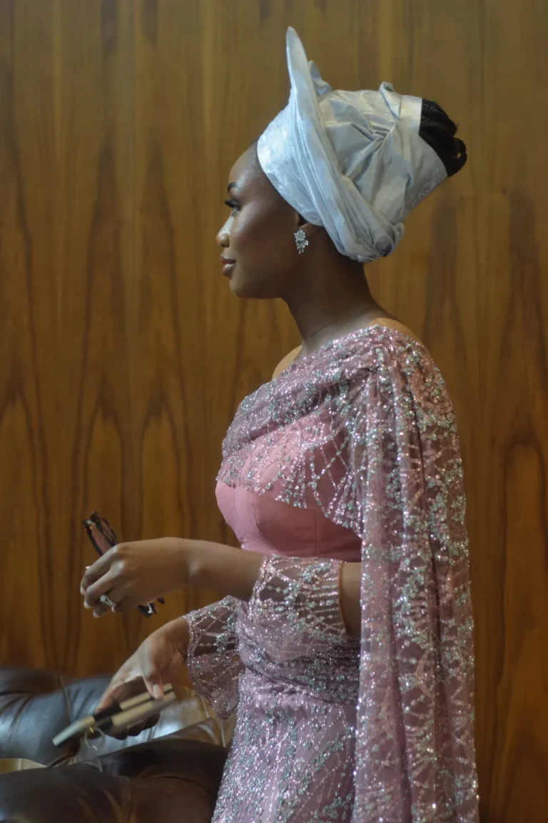 Woman in side profile wears a white gele and pink beaded gown against wood panelling.