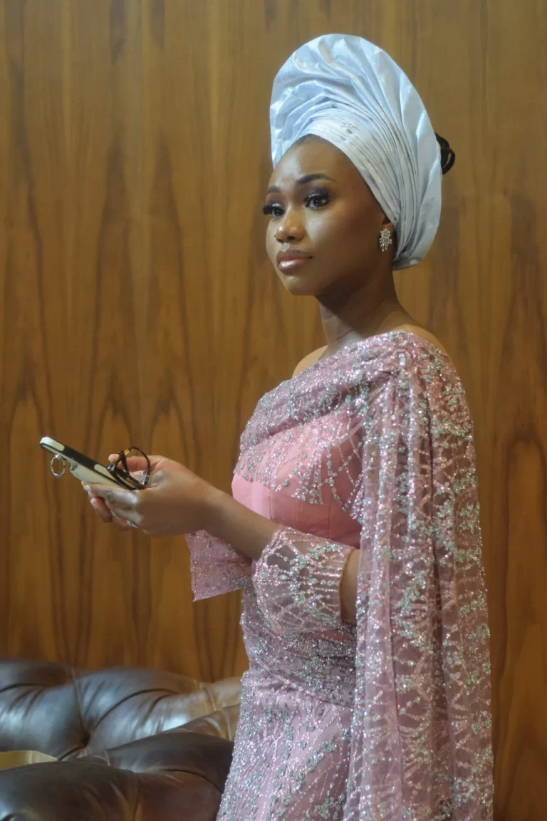 Woman in white gele and shimmering pink gown holds a pen and phone.
