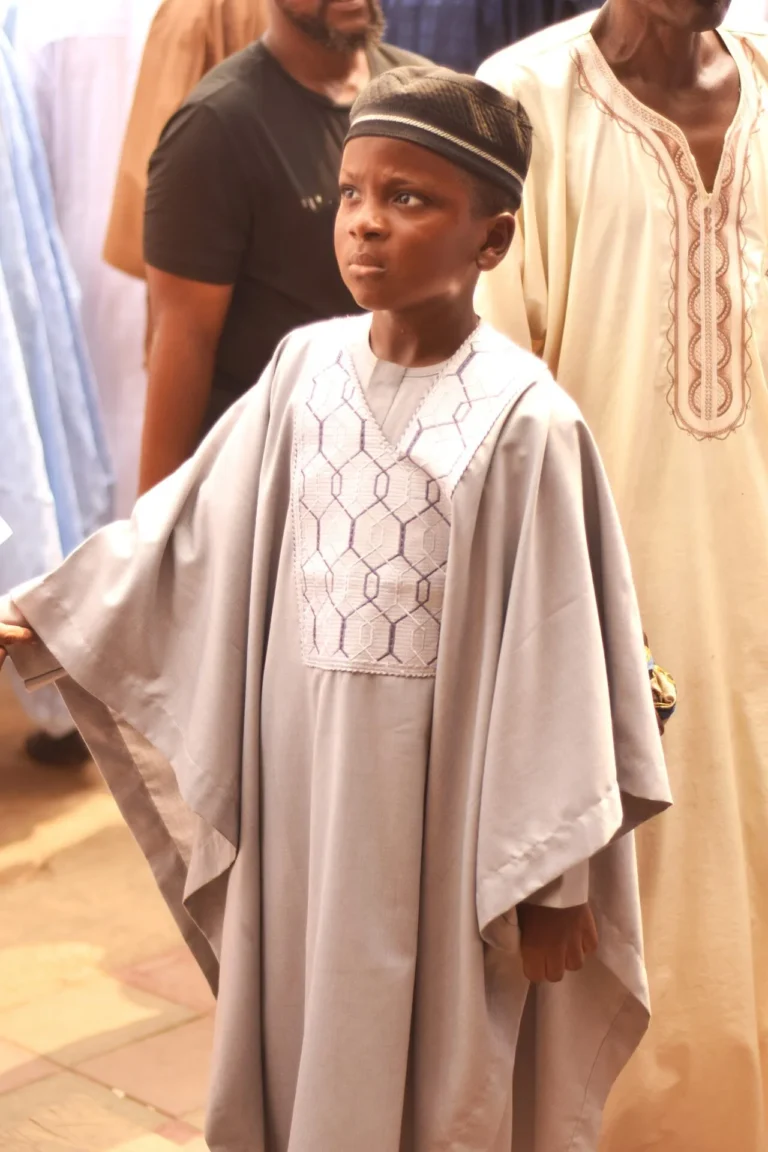 Young boy in pale grey agbada and embroidered cap stands among adult guests.