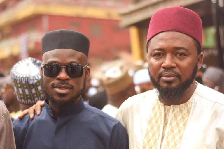 Two men pose closely, one in red fez, the other in sunglasses and dark robe.