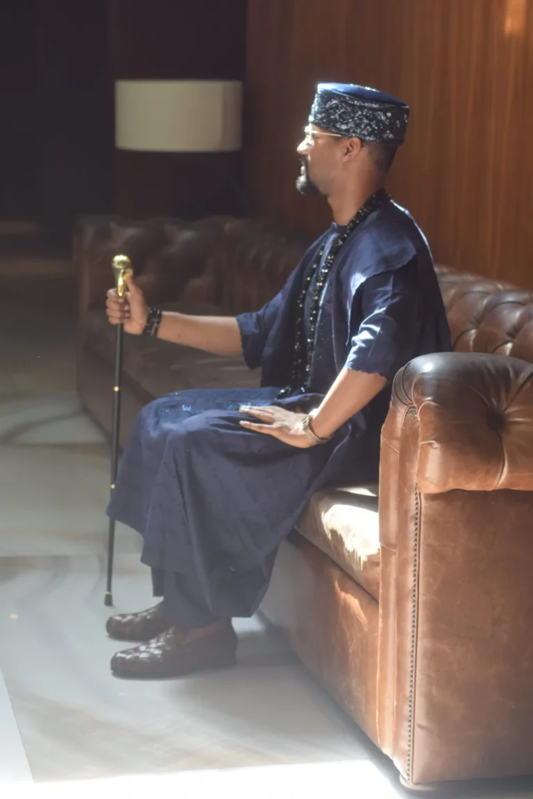 Man in navy agbada and cap sits on a brown leather sofa holding a cane.