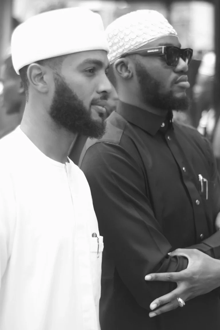 Two bearded men in white caps and dark shirt stand profile to camera.