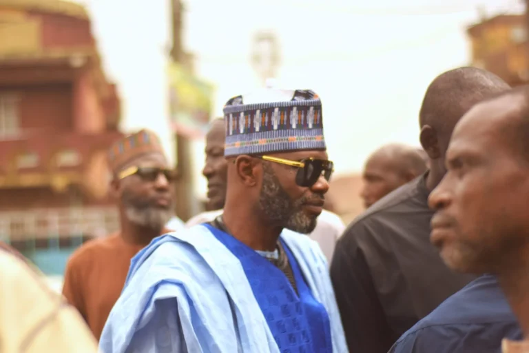 Man in blue agbada and patterned cap walks through a crowded street.