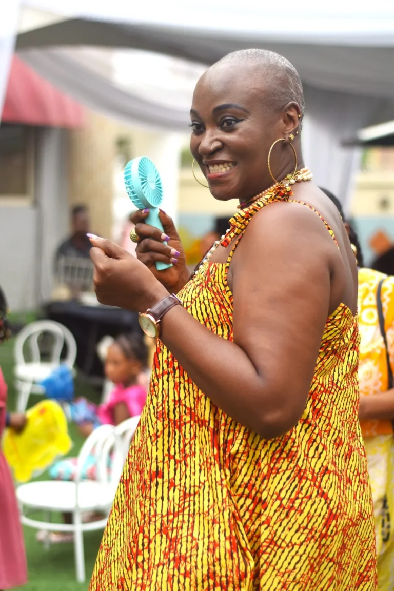 Woman in yellow and red African print dress laughs while holding a small turquoise handheld fan.