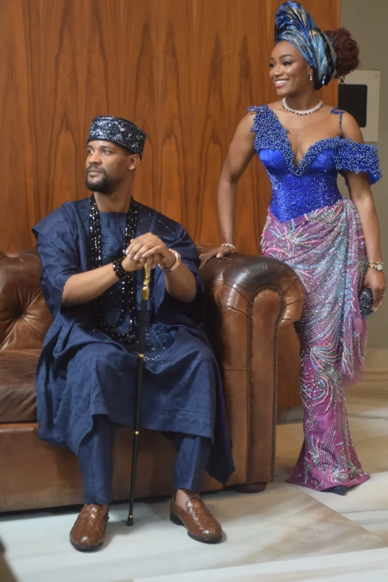 Couple in matching navy and royal-blue traditional Nigerian attire pose by a leather sofa.
