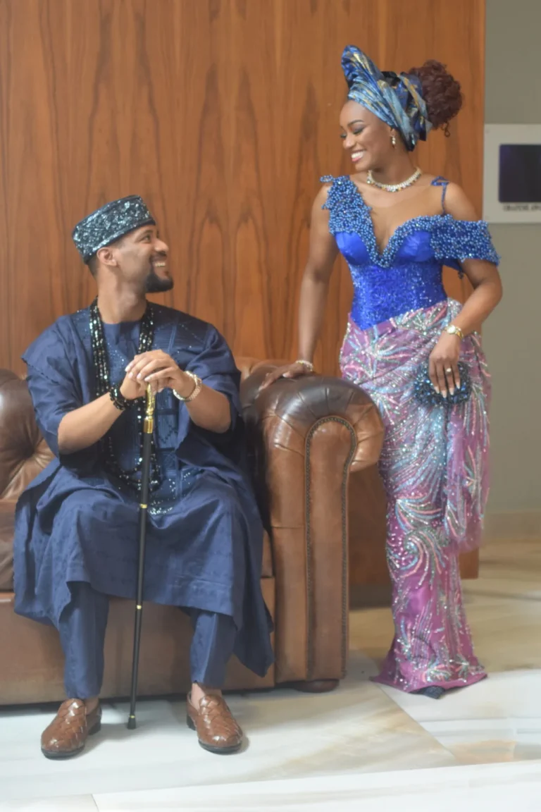 Bride in blue corset gown smiles down at seated groom in navy agbada with cane.
