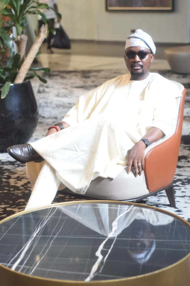 Man in cream agbada and sunglasses lounges in an orange armchair beside a marble table.