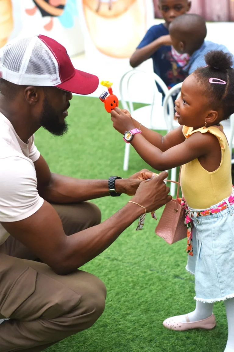 Father crouches to fasten a watch on his daughter's wrist as she plays with a toy.