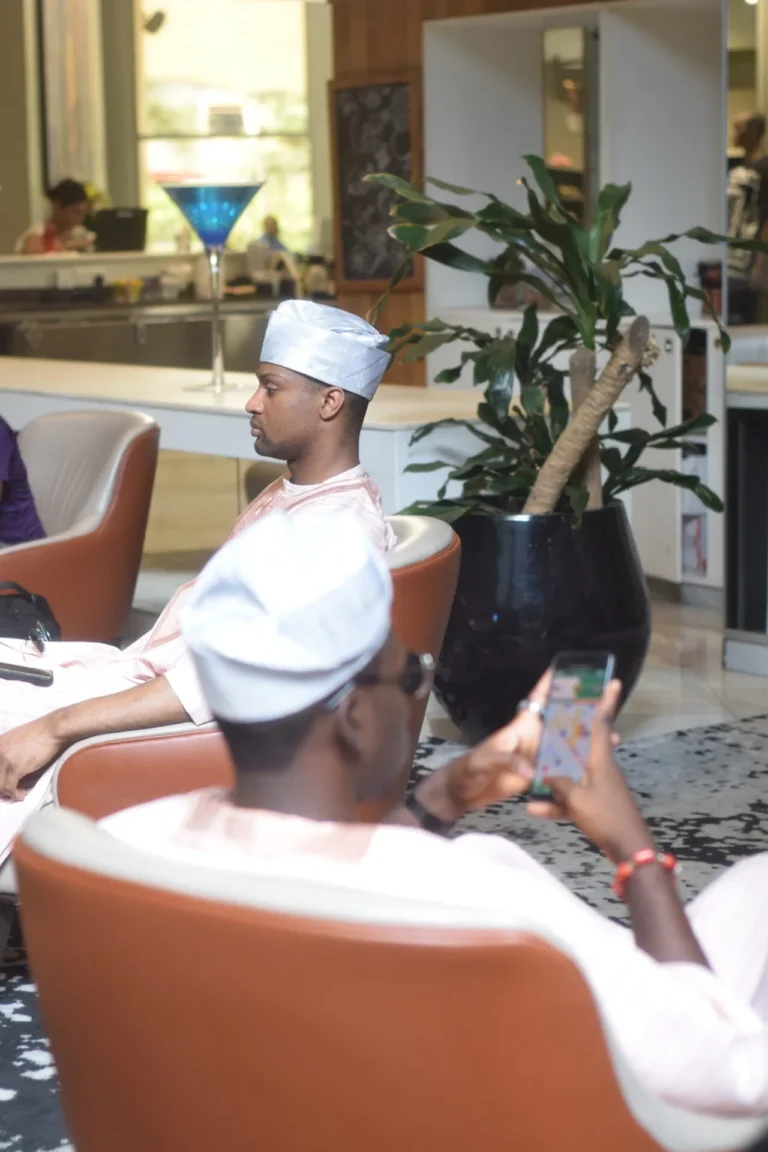 Two men in pale fila caps relax in lobby armchairs, one scrolling a phone.