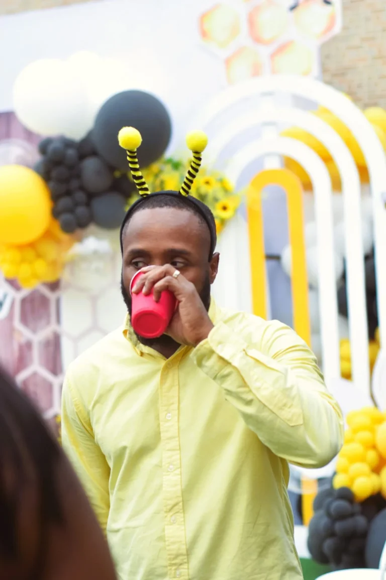 Man in yellow shirt and bee antennae drinks from a red plastic cup against a honeycomb backdrop.