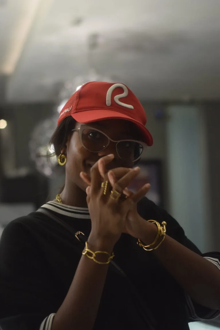 Person in red R cap and gold jewellery laughs behind crossed hands and rings.