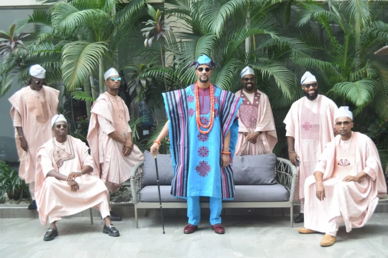 Groomsmen in dusty-pink agbadas flank a man in turquoise robes and red beads.