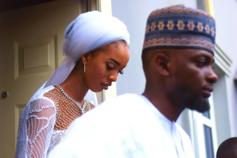 Bride in pearl gown and pale veil stands behind a man in patterned cap.