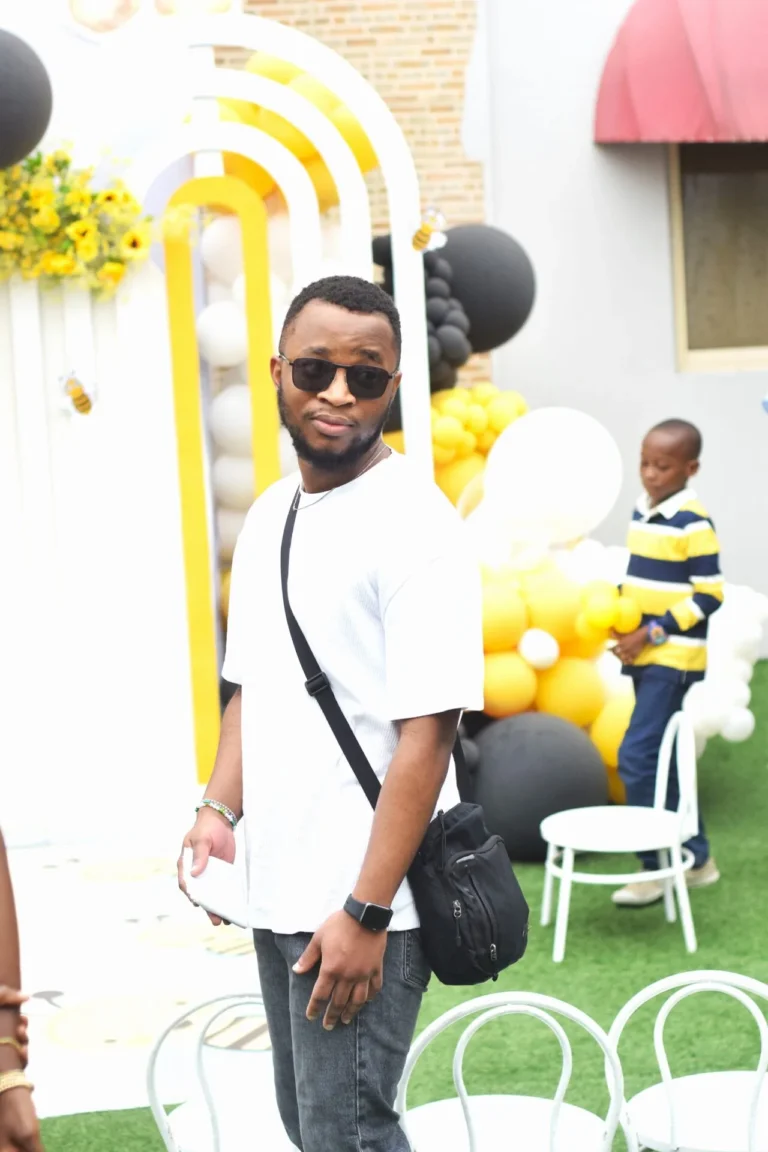 Man in sunglasses and white tee poses by yellow and black balloon arches at a party.