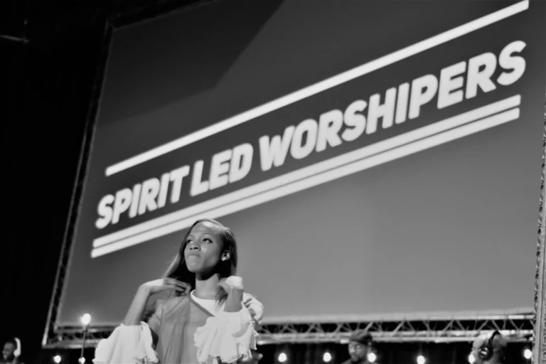 Black and white photo of woman on stage in ruffled dress beneath a 'Spirit Led Worshipers' screen.