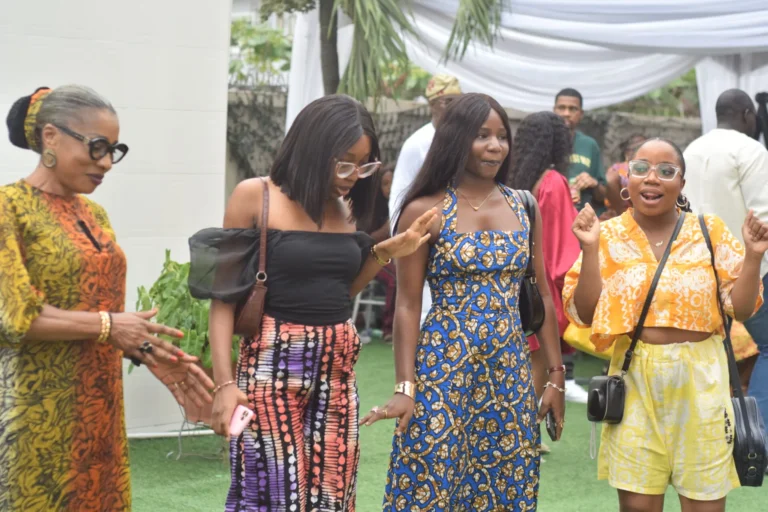 Four women in colourful patterned dresses chat and laugh at an outdoor celebration under a marquee.