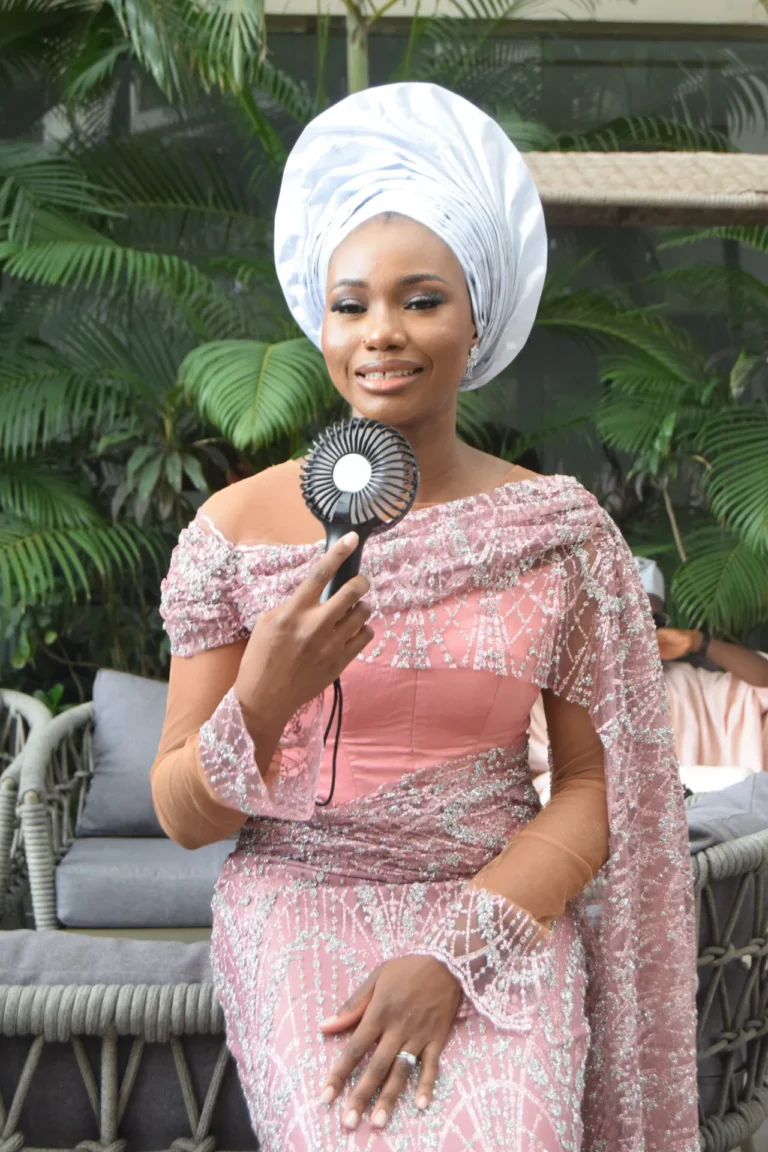 Woman in white gele and pink beaded gown holds a small hand-held fan outdoors.