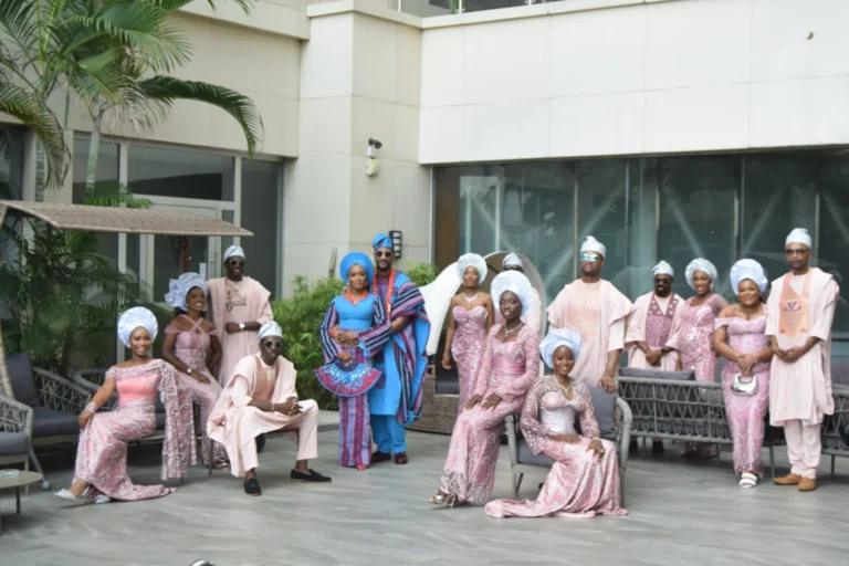 Large bridal party in coordinated dusty-pink and blue Nigerian wedding attire pose together.